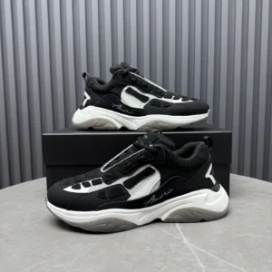 AMIRI Trainer sneakers in black and white, featuring a stylish design with prominent branding, displayed on a gray surface next to their box.