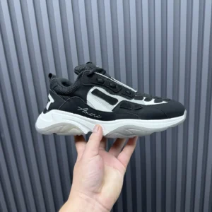 AMIRI Trainer black sneaker held in hand, featuring sleek design and contrasting white accents, showcased against a textured gray background.