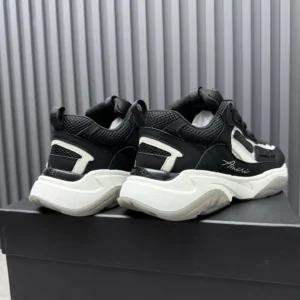 AMIRI Trainer sneakers in black and white, showcasing a stylish design with a prominent logo, displayed on a black box against a textured background.