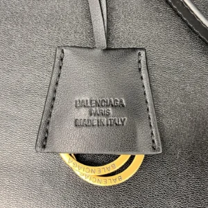 Balenciaga logo tag on Small Mailman Crossbody Bag, featuring embossed text "BALENCIAGA PARIS MADE IN ITALY" and a circular gold accent, showcasing luxury craftsmanship and brand identity.