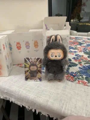 Pop Mart Labubu plush toy with bunny ears, accompanied by collectible card, displayed on a floral tablecloth amidst packaging boxes.