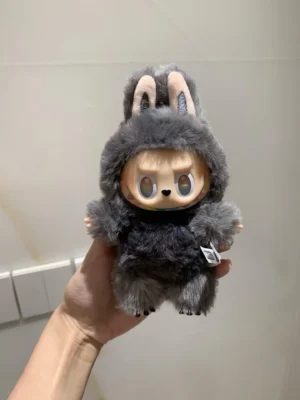 Pop Mart Labubu collectible doll in a gray rabbit costume, featuring large eyes and a fluffy texture, held in a hand against a neutral background.