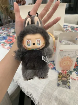 Hand holding Pop Mart Labubu plush toy in a gray bunny costume, showcasing colorful eyes and a playful design, with packaging visible in the background, emphasizing the collectible nature of the product.