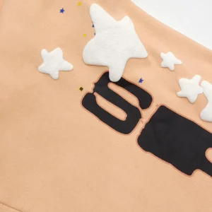 Sp5der Orange Pink Star Pants featuring a peach background with black lettering and white star appliqués, emphasizing unique fabric design and playful aesthetic.