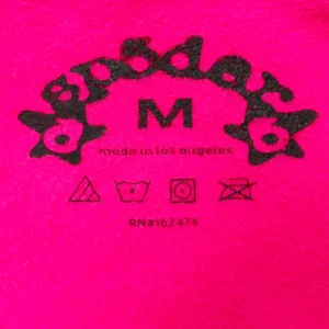 Label of SDER Rose Red Hot Diamond Black Word Legacy Hoodie featuring brand logo, size "M", and care instructions on vibrant pink fabric.