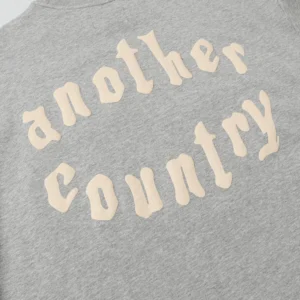 Gray hoodie featuring the text "another country" in a stylized font, showcasing the trendy design relevant to the Denim Tears brand.