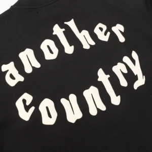 Black hoodie featuring the text "another country" in a bold, stylized font, showcasing trendy design elements relevant to Denim Tears fashion brand.