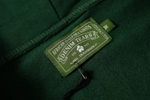 DENIM TEARS tag on green fabric, showcasing brand name and product details for the Cotton Flower hoodie.
