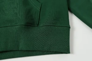 Close-up of a green hooded sweatshirt featuring a textured fabric and ribbed hem, representing the DENIM TEARS Cotton Flower product available for purchase.