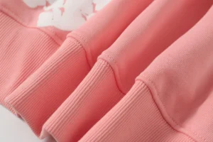 Cotton fabric details of DENIM TEARS hoodie in soft pink color, showcasing high-quality stitching and ribbed cuffs, emphasizing eco-friendly apparel design.