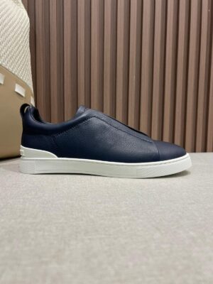 Minimalist navy leather slip-on shoes for men with a sleek design, featuring a white rubber sole and soft full-grain leather, set against a textured background.