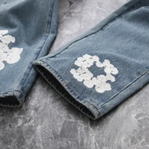 DENIM TEARS Water Washed Embroidery Vintage jeans featuring white floral embroidery on the cuffs, displayed on a textured gray surface.