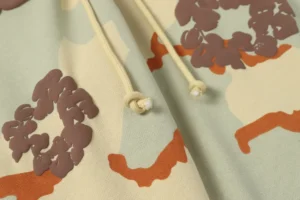 Close-up of DENIM TEARS cotton flower hair design on camouflage fabric, featuring drawstrings and vibrant colors, highlighting high-quality fashion details.