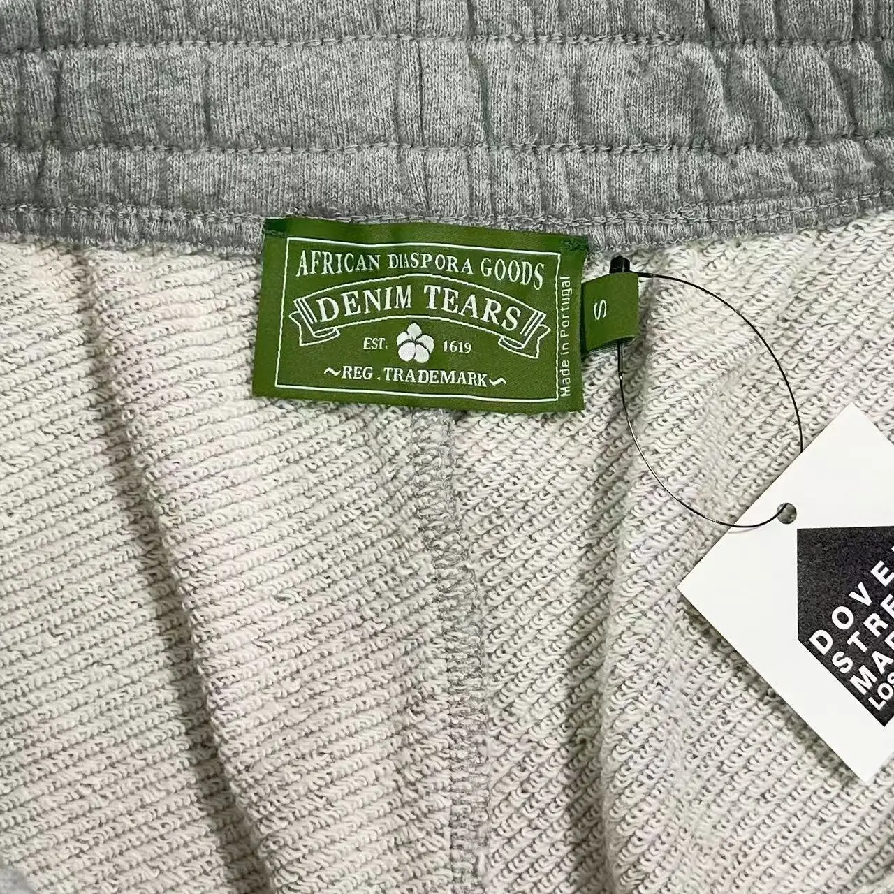 Denim Tears Main Line Hemp Grey Cotton pants label showing brand name, fabric details, and size tag in a textured fabric setting.