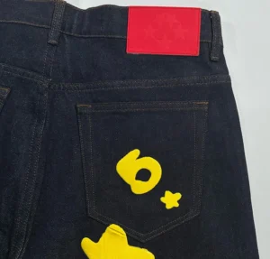 Sp5der Beluga Star Original Color Jeans featuring a dark denim fabric with yellow graphic designs on the back pocket and a red label at the waistband.