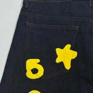 Sp5der Beluga Star Original Color Jeans featuring a dark denim fabric with vibrant yellow star and abstract designs on the back pocket.