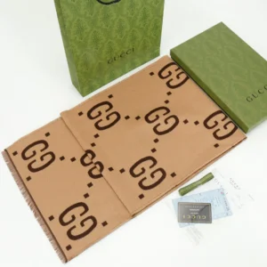 Gucci GG Large Logo Jacquard Wool Scarf displayed with packaging, featuring a double-sided design in tan and brown, alongside a green Gucci gift box and shopping bag.