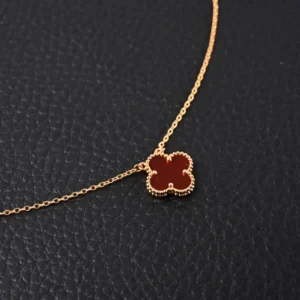 Van Cleef & Arpels Classic Clover Necklace in gold plating with red clover pendant on a textured black background.