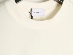 RHUDE branded tag on a cream-colored short-sleeved T-shirt, highlighting the clothing's premium quality for sale on an e-commerce fashion platform.