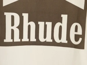 RHUDE retro logo print on a short-sleeved T-shirt, showcasing brand name in bold white letters against a brown background, emphasizing trendy streetwear fashion.