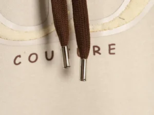 Supreme hoodie brown cotton drawstring with metal aglets on beige fabric background with "COUTURE" lettering